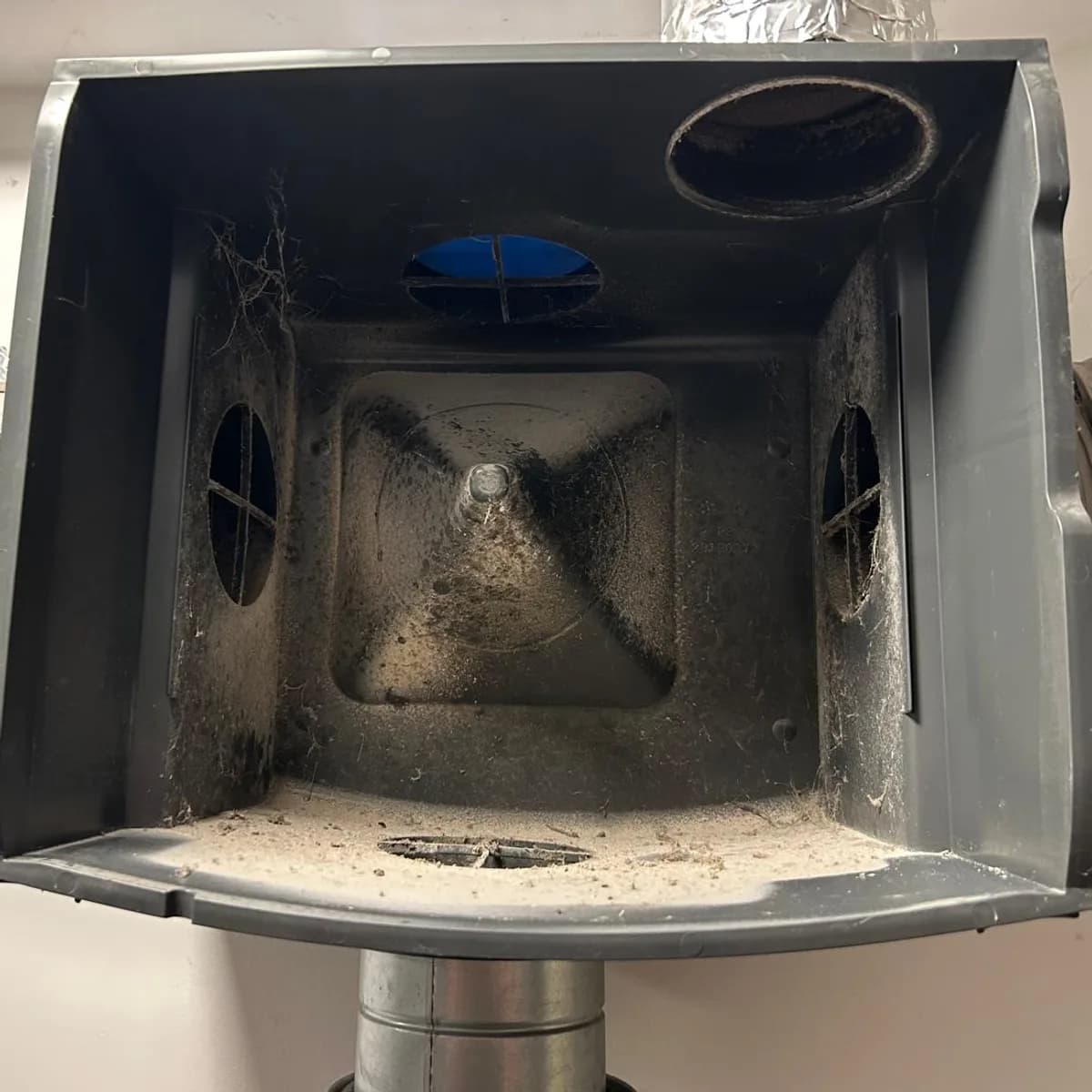 Caisson de VMC simple flux ouvert avant l'entretien : dépôt de poussière sur le fond et les parois, toiles d'araignée dans un coin.
